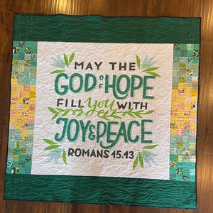 Könnte beinhalten: Eine weiße gesteppte Decke mit grünem Rand und grünem Blumendesign. Der Text "May the God of Hope fill you with Joy & Peace Romans 15:13" ist in Schwarz auf der Decke gedruckt.