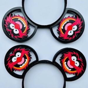 May include: Four black headbands with circular Animal character ears. The ears feature a red, pink, orange, and white design. The Animal character has a red nose, white teeth, and a wide smile. The headbands are on a white background.
