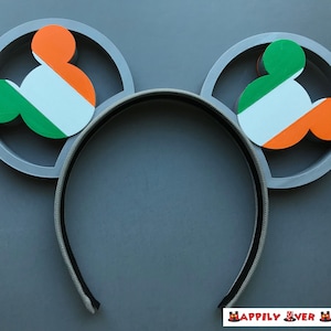 May include: Gray headband with two Mickey Mouse ear shapes, each featuring the Irish flag colors: green, white, and orange. The ears are set within gray circular frames. The headband is gray and black.