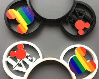 Op de afbeelding: Twee paar Mickey Mouse oren, een zwart en een wit. Het zwarte paar heeft een regenboog Mickey Mouse hoofd op het ene oor en een rood Mickey Mouse hoofd met het woord "LOVE" op het andere oor. Het witte paar heeft een regenboog Mickey Mouse hoofd op het ene oor en een rood Mickey Mouse hoofd met het woord "LOVE" op het andere oor.