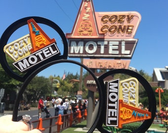 May include: A pair of black Mickey Mouse ears with a 3D Cozy Cone Motel sign attached to each ear. The sign is orange, yellow, and blue with the text "Cozy Cone Motel" in white. The sign is in front of a larger Cozy Cone Motel sign with the text "Cozy Cone Motel" in yellow and brown.