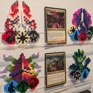 Pode incluir: Três cartas diferentes de Magic: The Gathering são exibidas em uma prateleira com três representações de pixel art correspondentes às criaturas das cartas. As cartas são "Kaalia of the Vast", "Animar, Soul of Elements" e "The Great Henge".