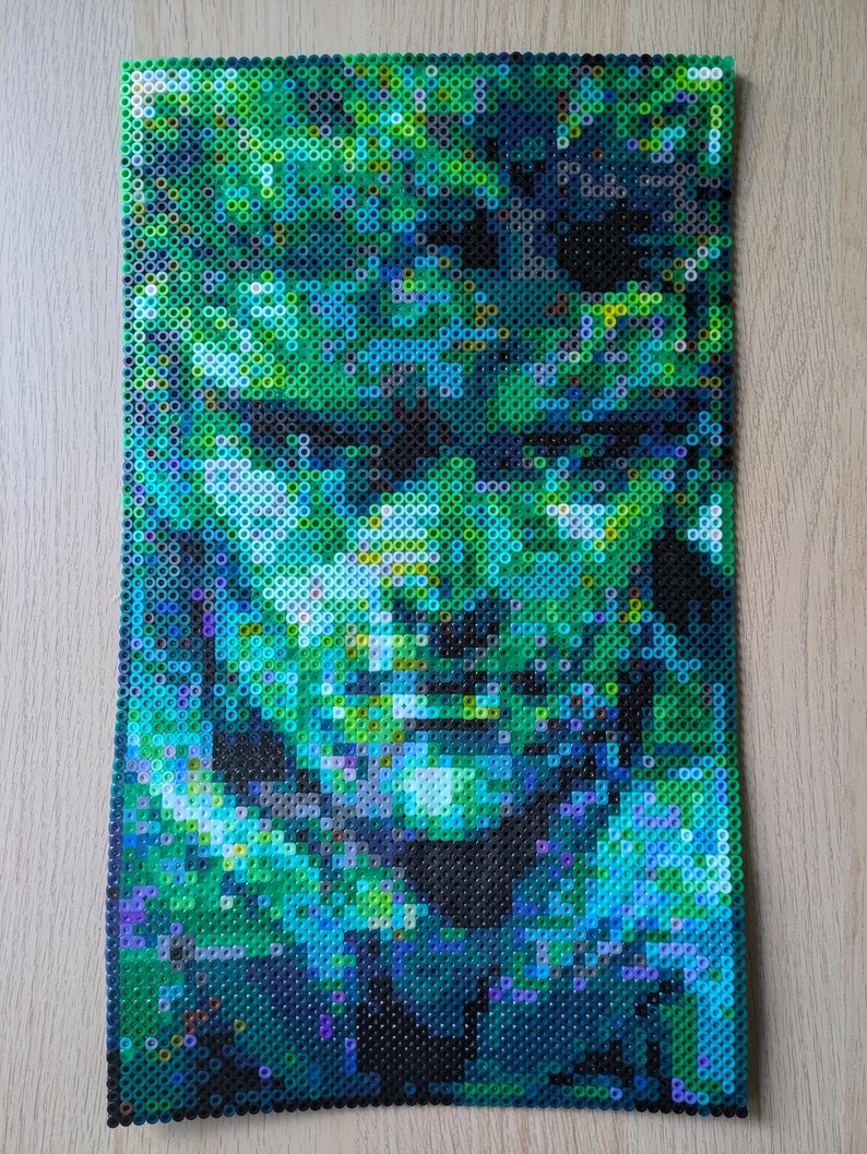 May include: A close-up of a pixel art image made with green, blue, and black beads. The image depicts a person's face.