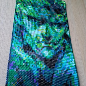 May include: A close-up of a green, blue, and black pixel art portrait of a person. The portrait is made of small, round beads.