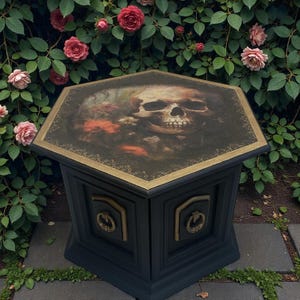 May include: A black hexagonal side table with a skull and floral design on the top. The table has gold trim and decorative accents. The table is set in a garden with pink roses and green foliage.