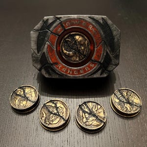 May include: A detailed image of a prop from the Power Rangers series. The prop includes a belt buckle with the words "Monster Rangers" and five matching coins. The buckle is gray and red, and the coins are gold and black.