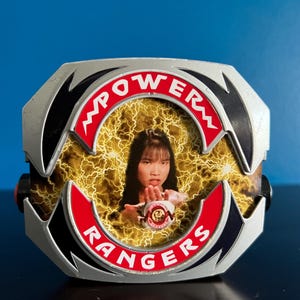 Peut inclure: Un morpher Power Rangers argenté et noir avec un centre rouge et jaune. Le morpher comporte les mots "POWER RANGERS" en lettres rouges et une photo d'une femme au centre.