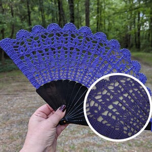 May include: A handmade, openwork crochet fan in a vibrant shade of blue. The fan features a scalloped edge and is mounted on a black, folding frame. The intricate crochet design is visible, showcasing the craftsmanship. The background is a blurred outdoor scene.