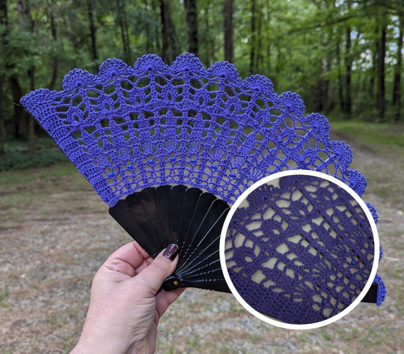 May include: A handmade, openwork crochet fan in a vibrant shade of blue. The fan features a scalloped edge and is mounted on a black, folding frame. The intricate crochet design is visible, showcasing the craftsmanship. The background is a blurred outdoor scene.