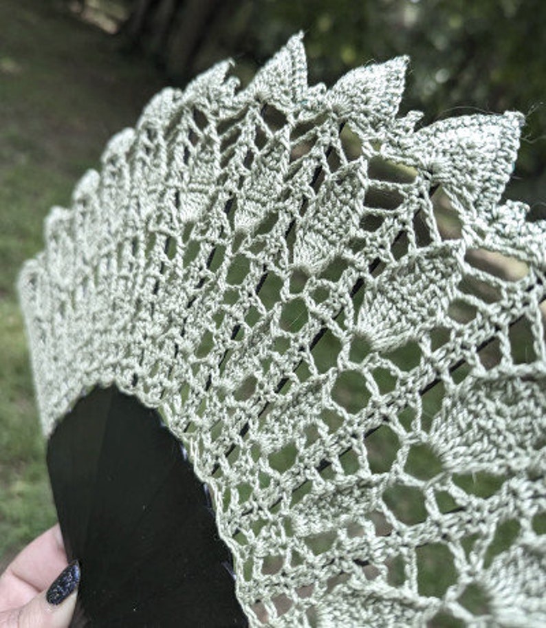 May include: A beige crocheted fan with a black handle. The fan is made of delicate lace and has a scalloped edge.