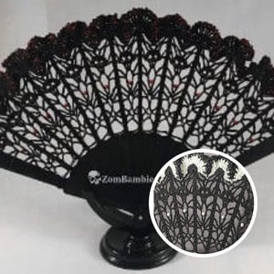 May include: Black lace hand fan with a decorative design. The fan features a black handle and a detailed openwork pattern. The top edge is trimmed with a dark red fringe. The fan is displayed on a black stand. The text "ZomBamble" is visible on the handle.
