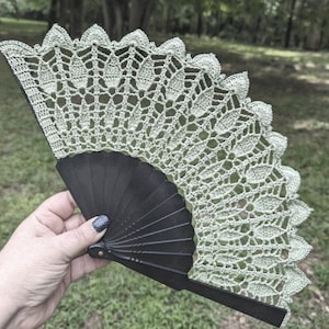May include: A green crocheted fan with a black folding handle. The fan is made of delicate lace and has a scalloped edge.