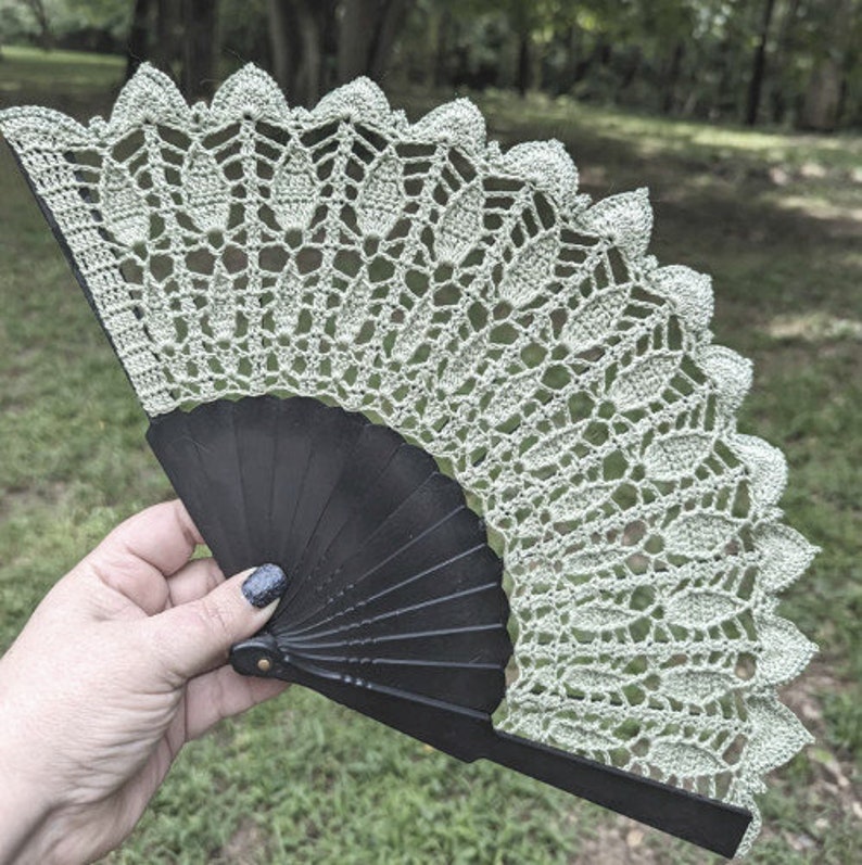 May include: A green crocheted fan with a black folding handle. The fan is made of delicate lace and has a scalloped edge.