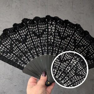 May include: A black, openwork crochet fan with a scalloped edge. The fan is held open, revealing the intricate pattern of the crochet. The handle is a dark gray color.
