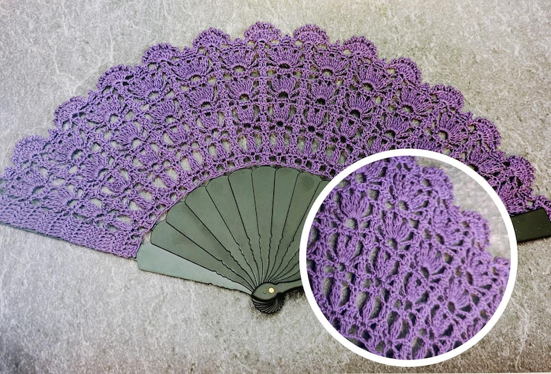 May include: A handcrafted, open purple crochet fan. The fan features a detailed, openwork design with a scalloped edge. The fan's base is a dark gray color. A circular inset shows a close-up of the crochet pattern.