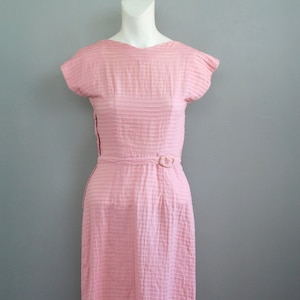 Könnte beinhalten: Hellrosa Kleid im Vintage-Stil mit einem strukturierten, horizontal gestreiften Muster. Das Kleid hat kurze Ärmel, einen V-Ausschnitt und einen passenden Gürtel mit kleiner Schnalle. Das Kleid wird auf einer Schaufensterpuppe präsentiert.