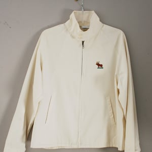 Puede incluir: Una chaqueta blanca con cremallera con un alce bordado en la parte delantera. La chaqueta tiene un cuello alto y dos bolsillos.