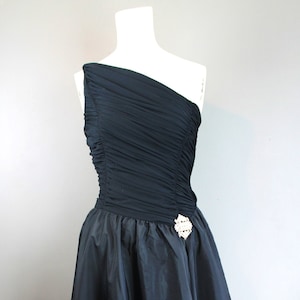May include: A black, one-shoulder dress on a mannequin. The bodice is ruched, and the skirt is a full A-line. A decorative brooch with white accents adorns the waist. The dress is a formal wear item.