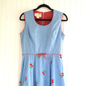 May include: A light blue sleeveless dress with a red polka dot lining and red cherry embroidery. The dress has a bow at the waist and is made of a lightweight fabric.