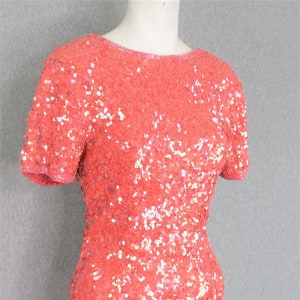 Peut inclure: Robe corail à paillettes avec manches courtes. La robe est recouverte de petites paillettes rondes qui captent la lumière, créant un effet scintillant. Elle a une encolure ronde et une silhouette ajustée.