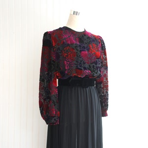 Peut inclure: Robe vintage sur mannequin. Le haut présente un motif floral rouge, rose et violet sur fond noir. Les manches longues et la taille sont noires. La jupe est en tissu noir plissé et transparent.