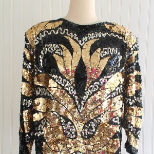 May include: A long-sleeved top covered in gold and black sequins. The design features a central gold pattern with black accents and a round neckline. The garment is displayed on a mannequin against a white background.