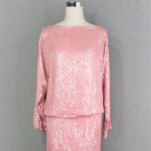 Op de afbeelding: Een roze pailletten tweedelig outfit, bestaande uit een top met lange mouwen en een knielange rok. De outfit wordt gepresenteerd op een mannequin tegen een grijze achtergrond. De pailletten creëren een glinsterend effect.