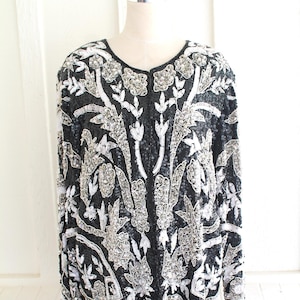May include: A black sequined jacket with a silver floral pattern. The jacket has a round neckline and long sleeves.