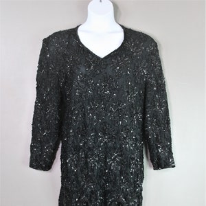 May include: Black sequined dress with a V-neck and long sleeves. The dress features a scalloped hemline with fringe detailing. The garment is displayed on a white mannequin, showcasing its design and texture. A vintage-inspired piece.
