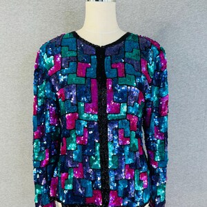 May include: A long-sleeve jacket covered in a geometric pattern of sequins in shades of magenta, teal, and dark blue. The garment features a round neckline and a vertical front opening trimmed with black beads, extending along the edges.