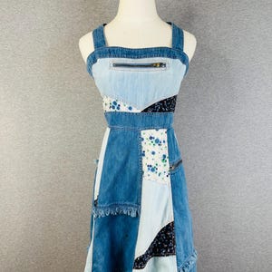 May include: A sleeveless patchwork dress constructed from various shades of blue denim and contrasting fabric panels. The design features a horizontal zipper pocket on the chest, a square neckline, and frayed hem details with floral and dark patterned accents.