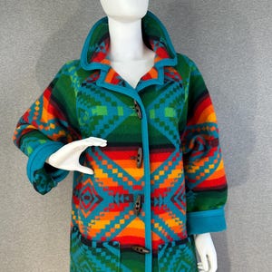 May include: A hooded coat featuring a vibrant geometric pattern in shades of teal, orange, red, and green. The coat has long sleeves, two front pockets, and a front closure secured by four dark brown toggle buttons with teal trim along the edges.