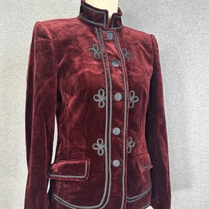 May include: A deep burgundy velvet jacket featuring a high collar, long sleeves, and a front button closure. The garment is accented with dark grey piping and decorative braided cord loops on the front panels and two flap pockets.