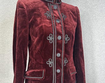 May include: A deep burgundy velvet jacket featuring a high collar, long sleeves, and a front button closure. The garment is accented with dark grey piping and decorative braided cord loops on the front panels and two flap pockets.