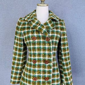 May include: A double-breasted pea coat featuring a textured houndstooth pattern in shades of olive green, teal, white, and brown. The coat has a wide notched collar, long sleeves, and six round brown buttons arranged in two vertical rows on the front.