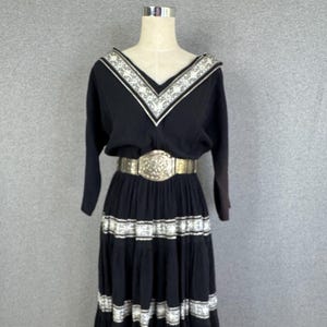 Pode incluir: Um vestido midi preto com decote em V com acabamento estampado em branco e preto, mangas três quartos e uma saia em camadas com faixas estampadas a condizer. Um cinto largo, metálico e em relevo está ajustado à cintura do vestido.