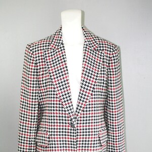 May include: A houndstooth blazer in black, red, and white, displayed on a mannequin. The jacket features a classic notched collar, two front pockets, and a single-button closure. The pattern is a repeating small-scale design.