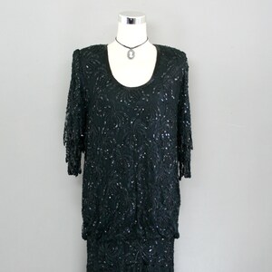 Peut inclure: Une robe noire à paillettes avec un col rond et une jupe fluide. La robe est sur un mannequin et porte un collier argenté avec un pendentif camée.