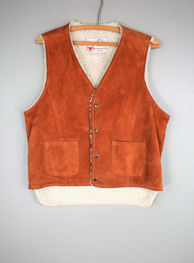May include: A brown suede vest with a white sherpa lining. The vest has a button closure and two pockets. The label on the vest reads "The Leather Shop".