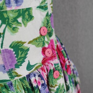 K&ouml;nnte beinhalten: Ein wei&szlig;es Kleid mit Blumenmuster in Rosa, Lila und Gr&uuml;n. Das Kleid hat einen Knopfverschluss vorne und einen R&uuml;schenrock.