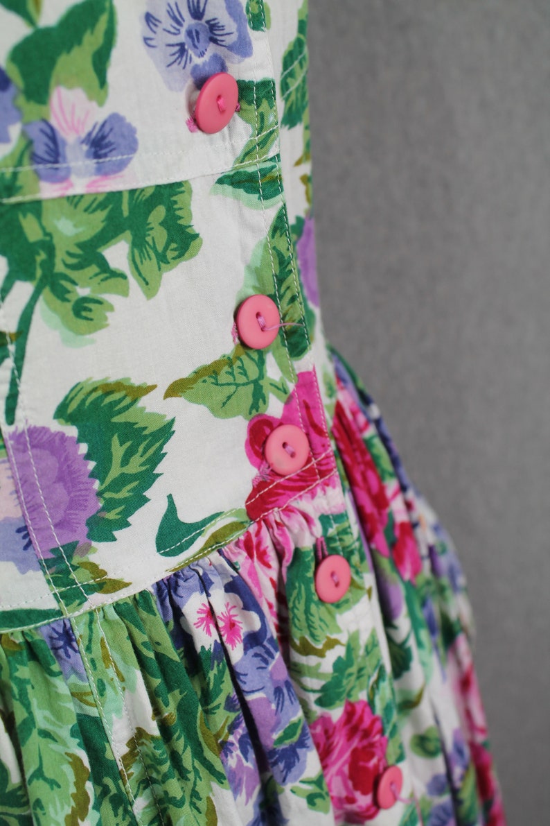 K&ouml;nnte beinhalten: Ein wei&szlig;es Kleid mit Blumenmuster in Rosa, Lila und Gr&uuml;n. Das Kleid hat einen Knopfverschluss vorne und einen R&uuml;schenrock.