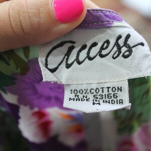 K&ouml;nnte beinhalten: Ein wei&szlig;es Etikett mit schwarzem Text, auf dem "Access" und "100% Baumwolle R.N. 53166 Hergestellt in Indien" steht, auf einem gr&uuml;n-lila-wei&szlig;en Blumenstoff.