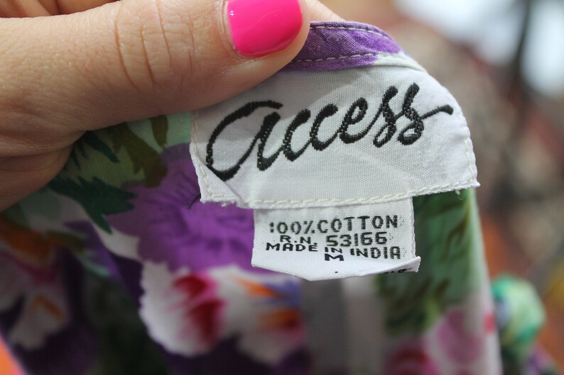 K&ouml;nnte beinhalten: Ein wei&szlig;es Etikett mit schwarzem Text, auf dem "Access" und "100% Baumwolle R.N. 53166 Hergestellt in Indien" steht, auf einem gr&uuml;n-lila-wei&szlig;en Blumenstoff.
