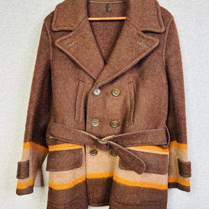 May include: A brown double-breasted wool coat featuring a wide collar, long sleeves with cuffs, and a matching belt at the waist. The lower section of the coat displays horizontal stripes in shades of tan and orange, with two large pockets.