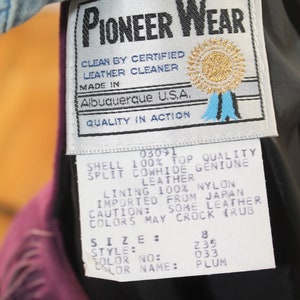 May include: A white tag with black text that reads "Pioneer Wear" and details about the leather jacket. The tag also includes the text "Clean by Certified Leather Cleaner", "Made in Albuquerque U.S.A.", and "Quality in Action". The tag also includes a gold star with a blue ribbon. The tag is attached to a purple leather jacket.