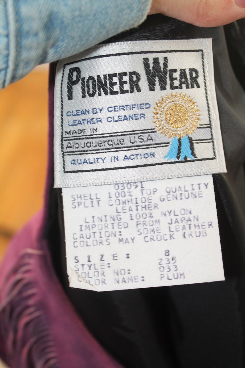 May include: A white tag with black text that reads "Pioneer Wear" and details about the leather jacket. The tag also includes the text "Clean by Certified Leather Cleaner", "Made in Albuquerque U.S.A.", and "Quality in Action". The tag also includes a gold star with a blue ribbon. The tag is attached to a purple leather jacket.