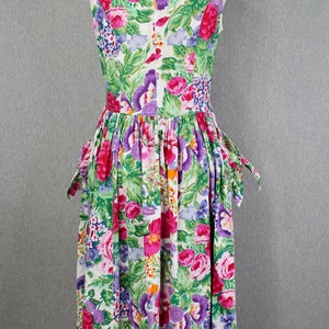 K&ouml;nnte beinhalten: Ein wei&szlig;es, &auml;rmelloses Kleid mit einem Blumendruck mit rosa, lila und gr&uuml;nen Blumen. Das Kleid hat einen taillierten Oberteil und einen vollen Rock mit Taschen.