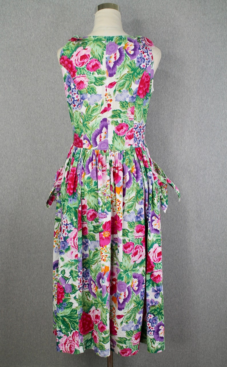 K&ouml;nnte beinhalten: Ein wei&szlig;es, &auml;rmelloses Kleid mit einem Blumendruck mit rosa, lila und gr&uuml;nen Blumen. Das Kleid hat einen taillierten Oberteil und einen vollen Rock mit Taschen.