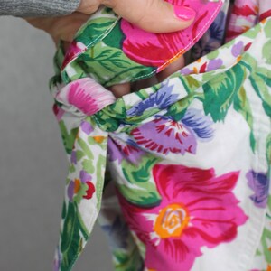 K&ouml;nnte beinhalten: Ein wei&szlig;er Stoff mit einem Blumenmuster in den Farben Rosa, Lila, Gr&uuml;n und Rot. Der Stoff hat oben eine Schleife und ist zum Erstellen einer Tasche umgeklappt.