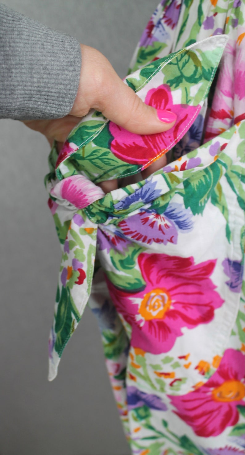 K&ouml;nnte beinhalten: Ein wei&szlig;er Stoff mit einem Blumenmuster in den Farben Rosa, Lila, Gr&uuml;n und Rot. Der Stoff hat oben eine Schleife und ist zum Erstellen einer Tasche umgeklappt.
